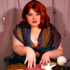 Lucrezia is a red-haired woman with sideswept bangs, loose curls. She has a multicolored scarf over her shoulders and a white tank top under it. She leans towards the viewer with one hand pushing a key towards them. The table is cluttered with other items.