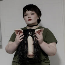 Amanda has a dark boycut with straight bangs. She wears a choker and army green shirt. Held over her chest in hands which have faint stains under the nails is a (latex) pig head mask with long dark hair.