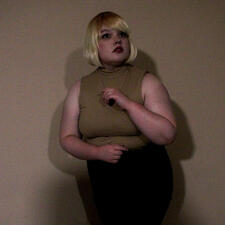 Ashley Graham (RE4R.) Her blonde bob has brown roots, and she seems less fearful than the previous picture. She has a tan turtleneck with no sleeves while the lower half of the body is too dark to tell. She has a necklace. Her arms are held over her torso.
