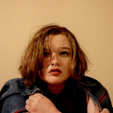 Centered low in the picture on a yellow backdrop is a girl. Her straight brown hair is disheveled, and she appears panicked. She holds a jean jacket around her shoulders. The jacket has red piping around the zippers. Her hands are covered in fake dirt.