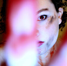 A hand blocks most of the screen, reaching out for the viewer. Beyond that is a face covered in fake blood and dirt. The person has short, curly, brown hair cropped around the ears and looks directly at the viewer with a single red eye visible.