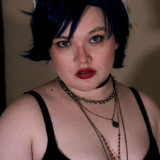 The subject has short, dark blue hair that flips up on their right side. Their eyes are a saturated blue, and their lipstick is dark and smudged. Around their neck is a set of layered necklaces, and they wear a black tank top.