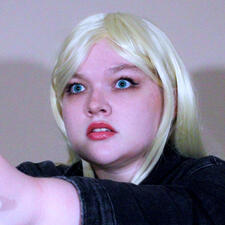 She has pale blonde hair with the bangs pinned out of her face. Her blue eyes are wide in mock-horror. She wears a black jean jacket, and her hands are held in front of her as though holding a gun, but it is offscreen.