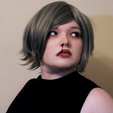 Seven has gray hair that is in a short Karen-ish bob and swept out of her face. She has on a black, sleeveless, mock-neck top. Fake blood trails from her right nostril and over her lips. She looks off towards the right of the picture.