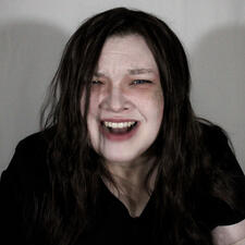 Amy Katz has long dark hair and a bruise on her left cheek. She looks tired and is wearing plain black scrubs. Her face is twisted into a scream.