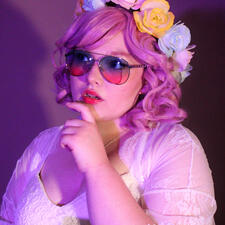 A person with a pink curly bob, a flower crown with alternating pink/yellow/blue flowers, and gradient aviators that are blue at the top, pink at the bottom. They have a lacy white top/coverup. There is a chain around their neck and one hand up.